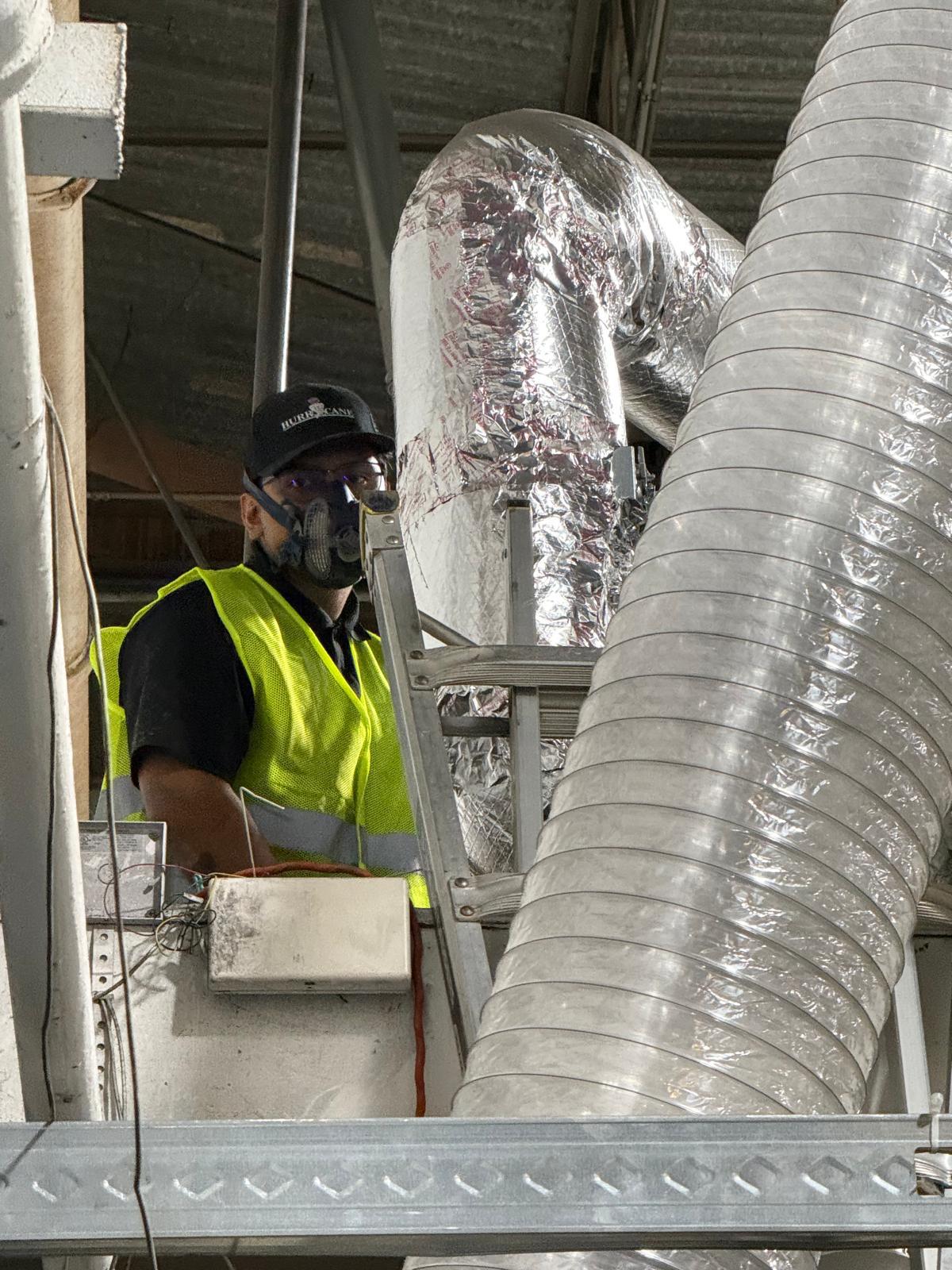 Factory ceiling work — technician in respirator servicing insulated supply duct above structural beams